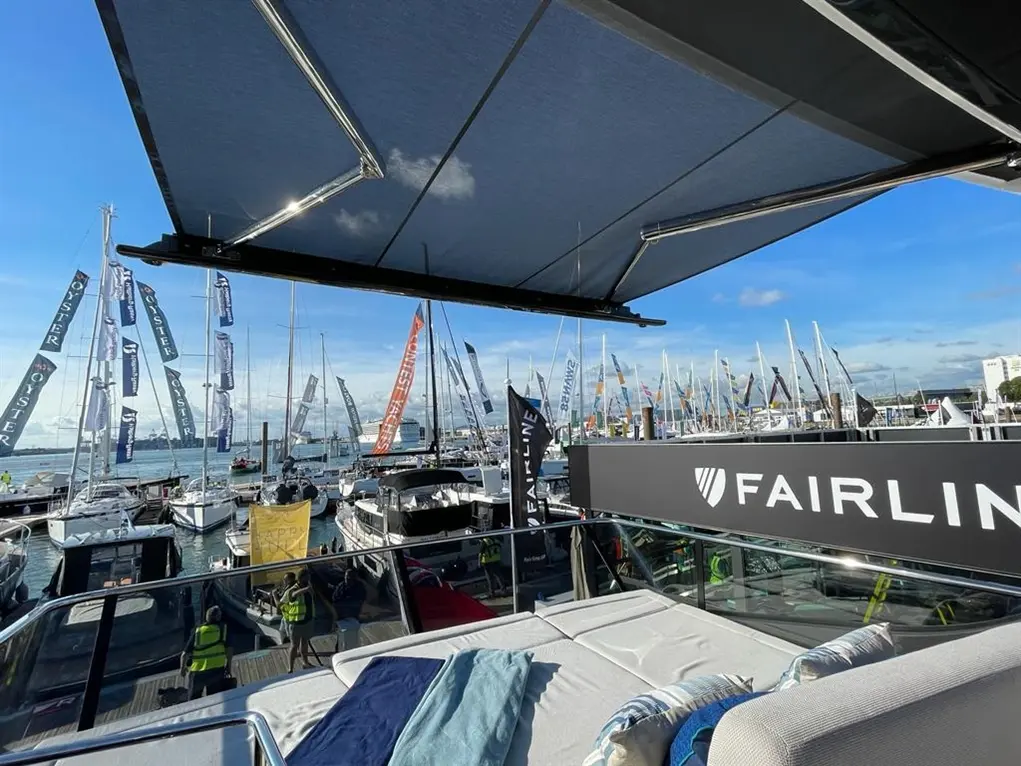 View from a yacht with a sunshade, showcasing numerous boats and flags at a boat show, featuring the Fairline logo prominently.